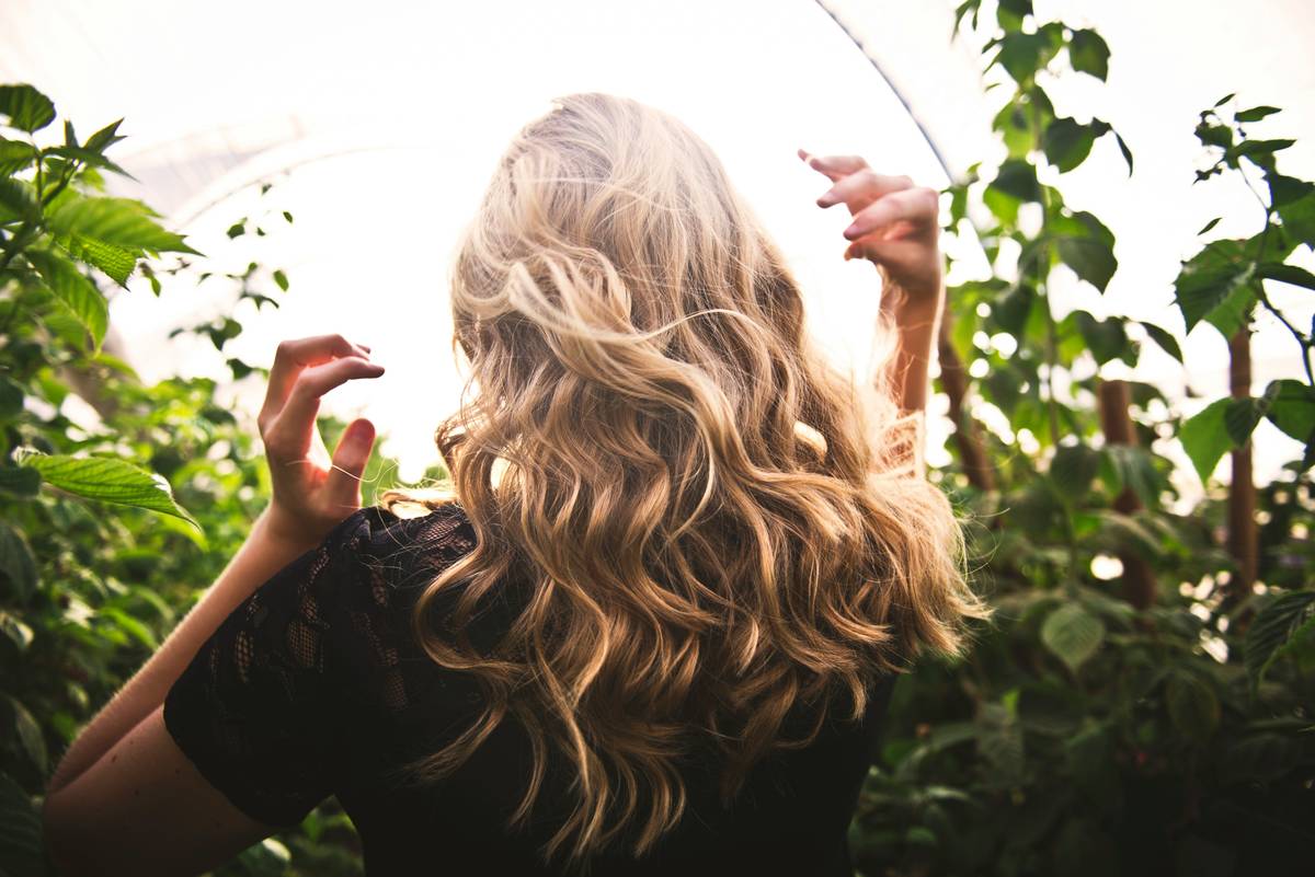 Woman applying summer hair conditioner to damp mid-length hair under shaded outdoor patio, visible UV-protective spray mist nearby